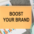 Picture of a notebook on a desk. There's a sentece written on the hard cover of the notepad that reads 'boost your brand'